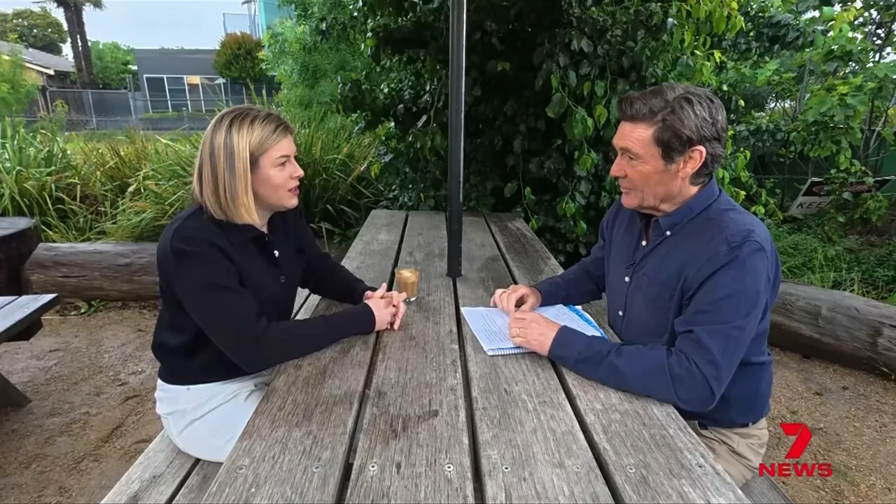 Two-shot of Jess Wilson and an interviewer seated across a wooden table, engaged in conversation outdoors