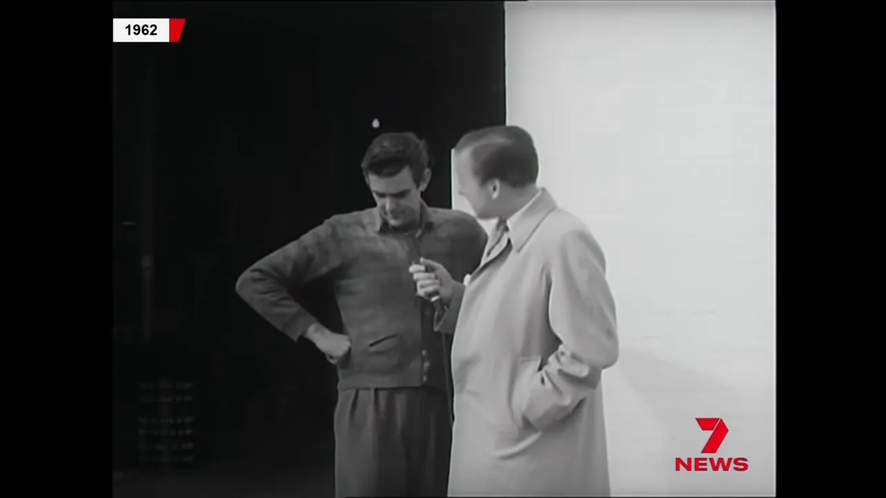 1962 black-and-white screenshot of a TV presenter holding a microphone while speaking to a man on his doorstep