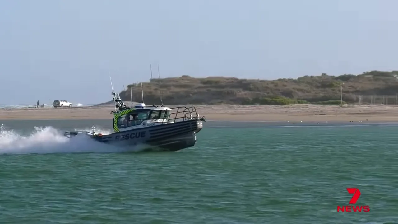 Close side view of a rescue boat cutting through the water with spray, sand dunes and breaking surf in the background.
