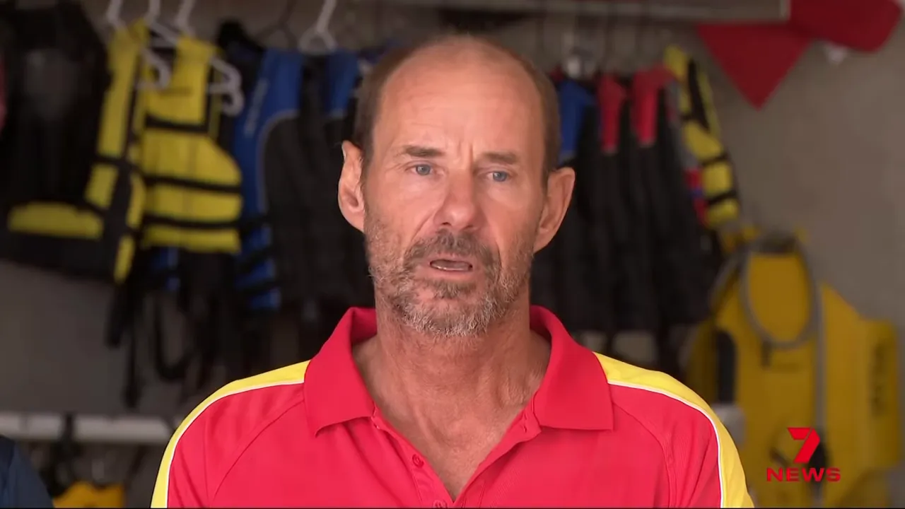 Frontal portrait of a surf lifesaver in a red rescue shirt with lifejackets and rescue gear visible behind him.