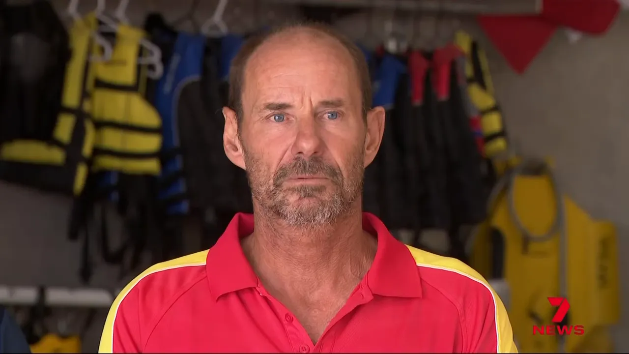 Frontal portrait of a surf lifesaver in a red rescue shirt with lifejackets hanging behind him.