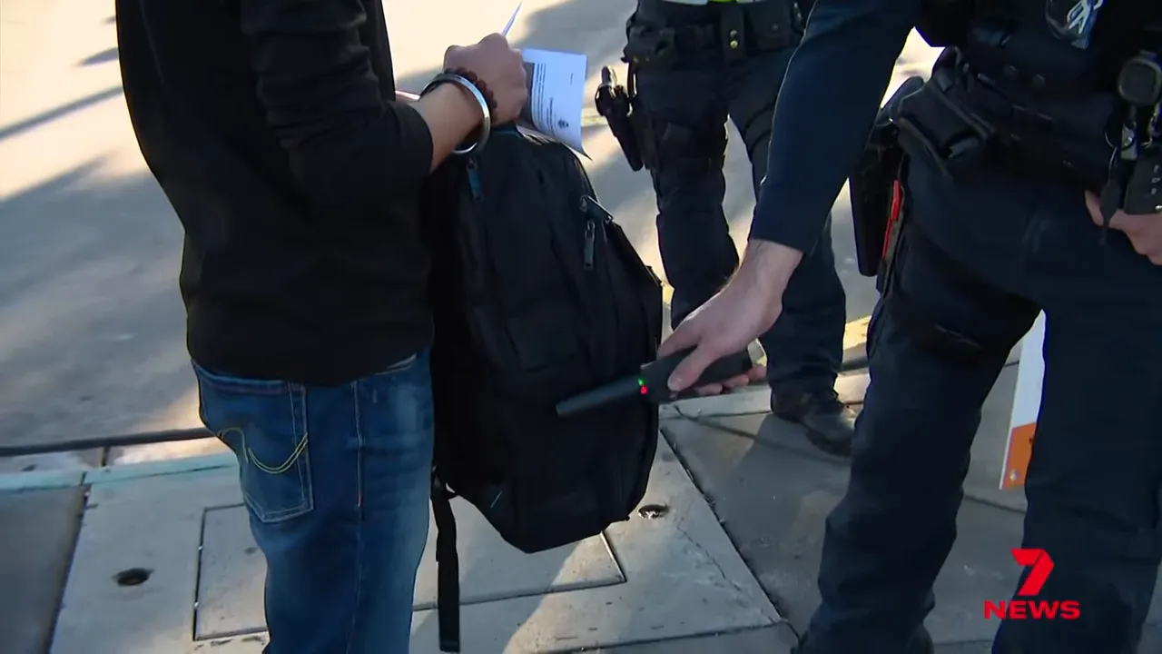 Close-up view of a police officer using a handheld wand scanner on a backpack during a routine search.