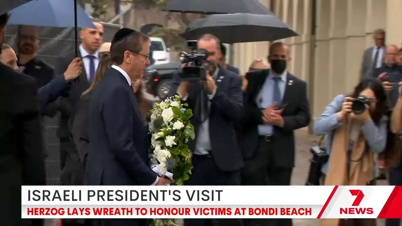 A dignitary in a suit holding a large floral wreath while photographers and aides stand under umbrellas
