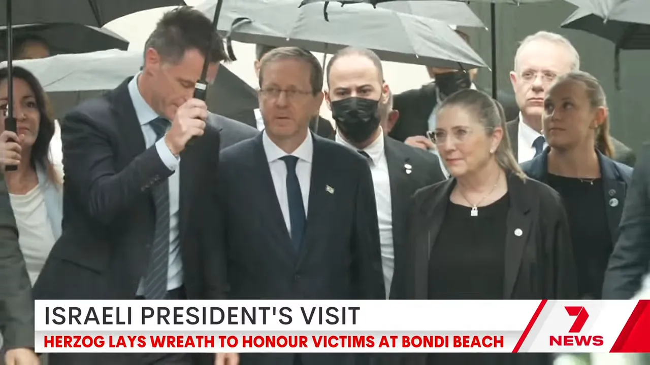 Group of officials and aides under umbrellas at a memorial with a news banner at the bottom of the frame.