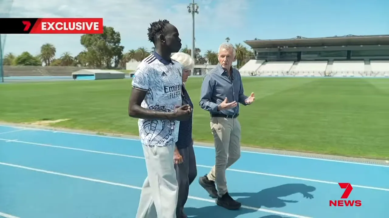 Sprinter and interviewers walking on a blue athletics track with the grandstand and grass field in the background.