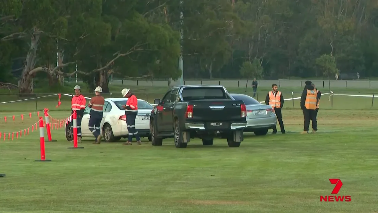 Workers and vehicles near a cordoned-off area on a sports oval after a sinkhole appeared