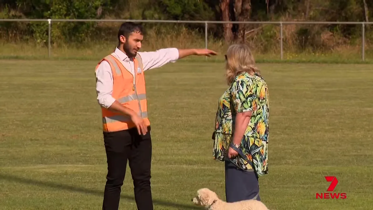 Man in an orange high-visibility vest gesturing with his arm while a woman with a small dog listens on a sports oval