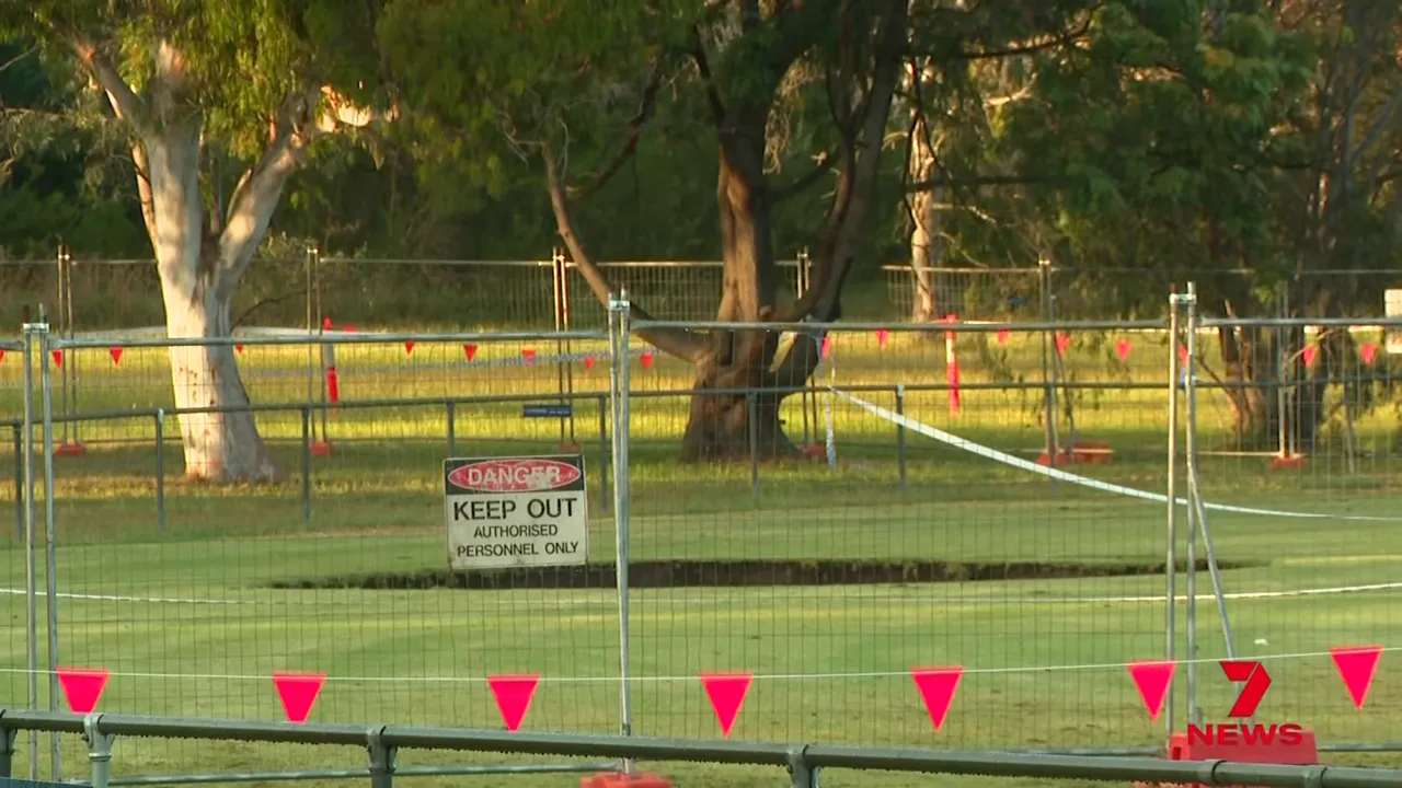 Wide shot of fenced sinkhole with Keep Out sign and exclusion tape on sports oval