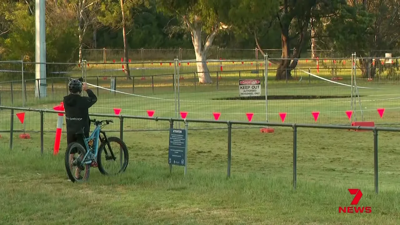 Person with bicycle observing fenced sinkhole on sports oval with warning flags