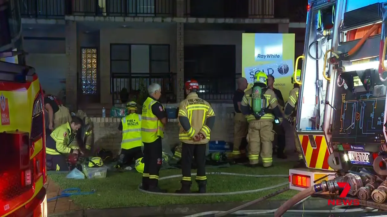 Firefighters and fire trucks at the front of a residential property at night with a Ray White sold sign on the lawn.
