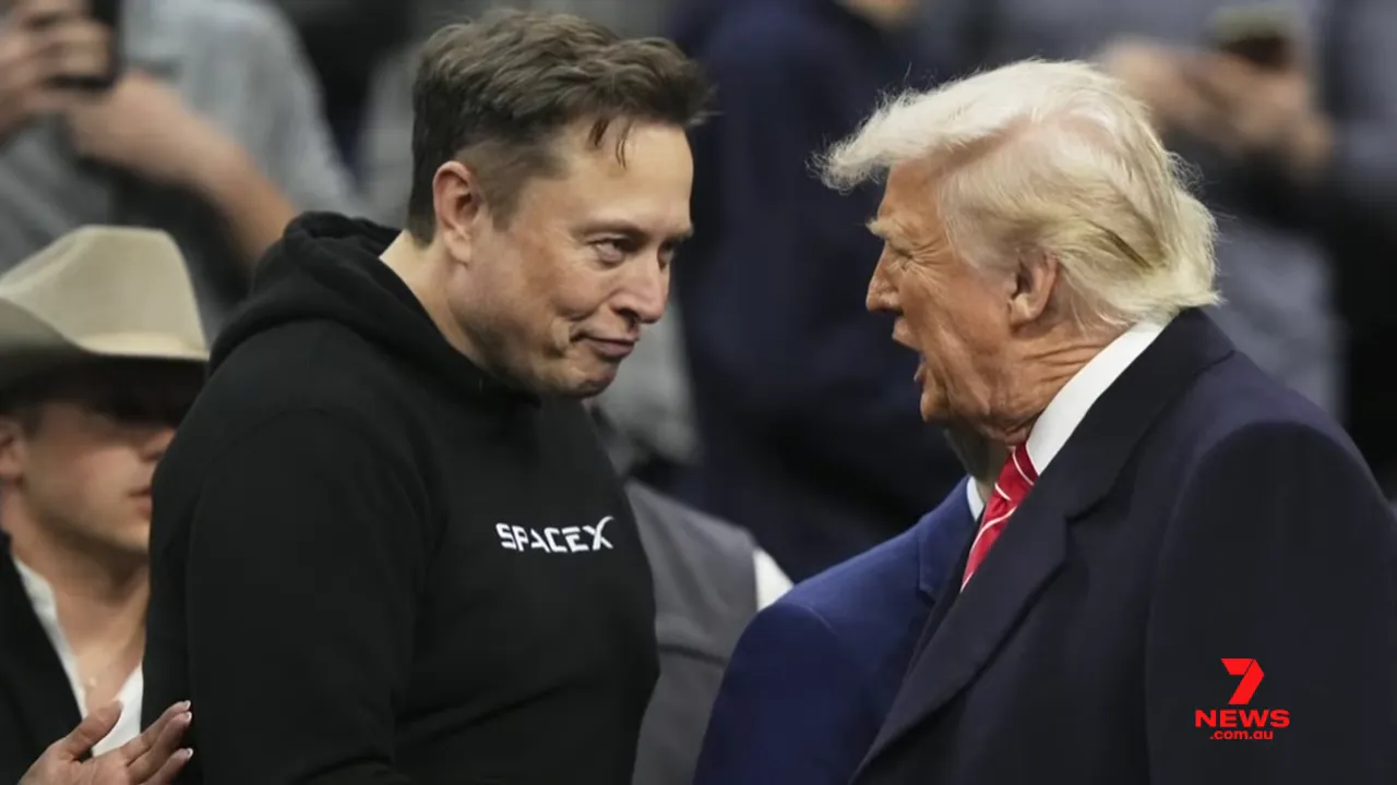 Two men facing each other closely at an event; one in a black hoodie with a visible logo, the other in a suit and red tie.