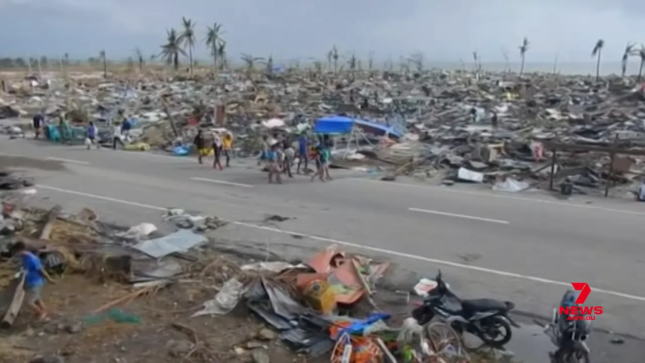 Wide, clear view of a road lined by flattened homes, scattered debris and stripped palm trees after a tropical cyclone.