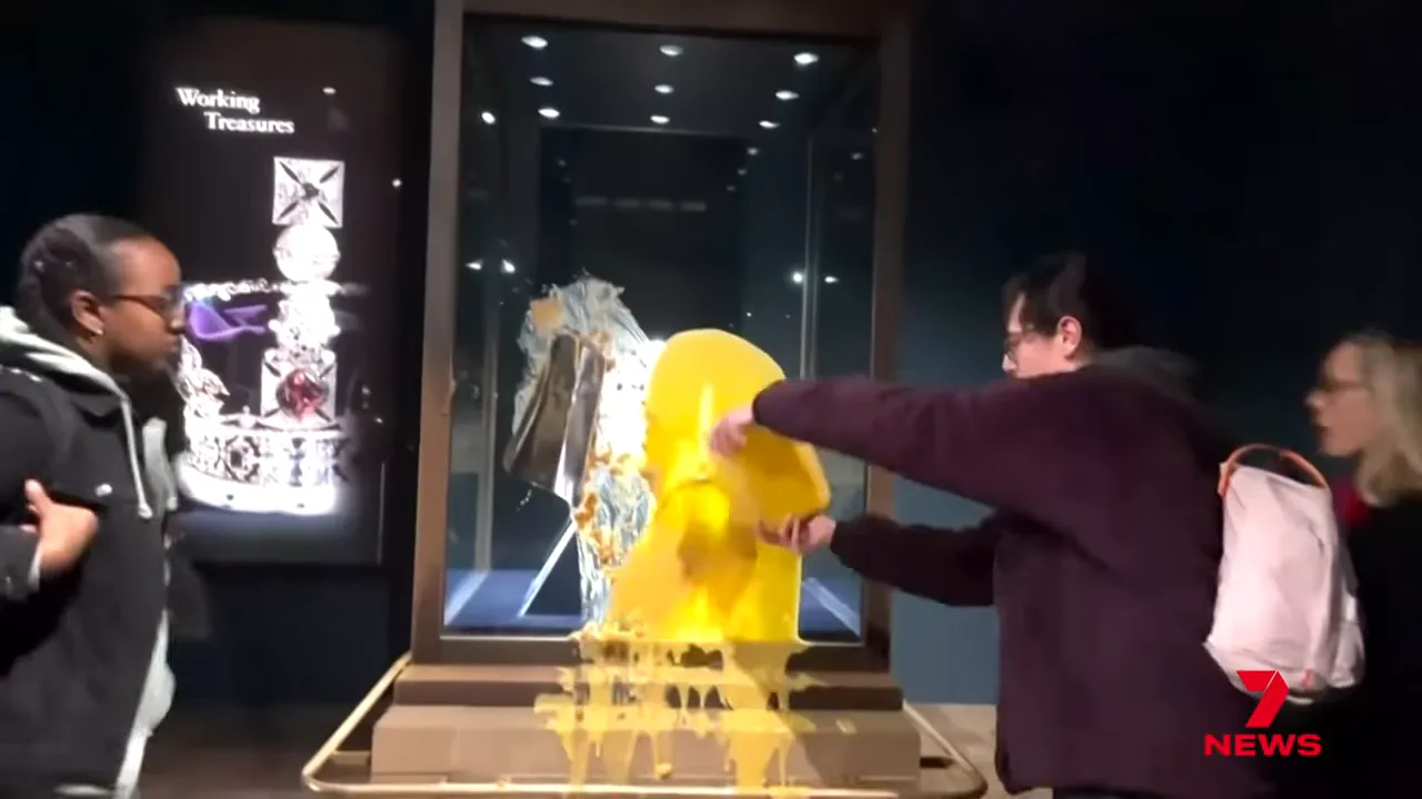 Person pouring yellow custard onto the glass case protecting the Imperial State Crown while visitors look on