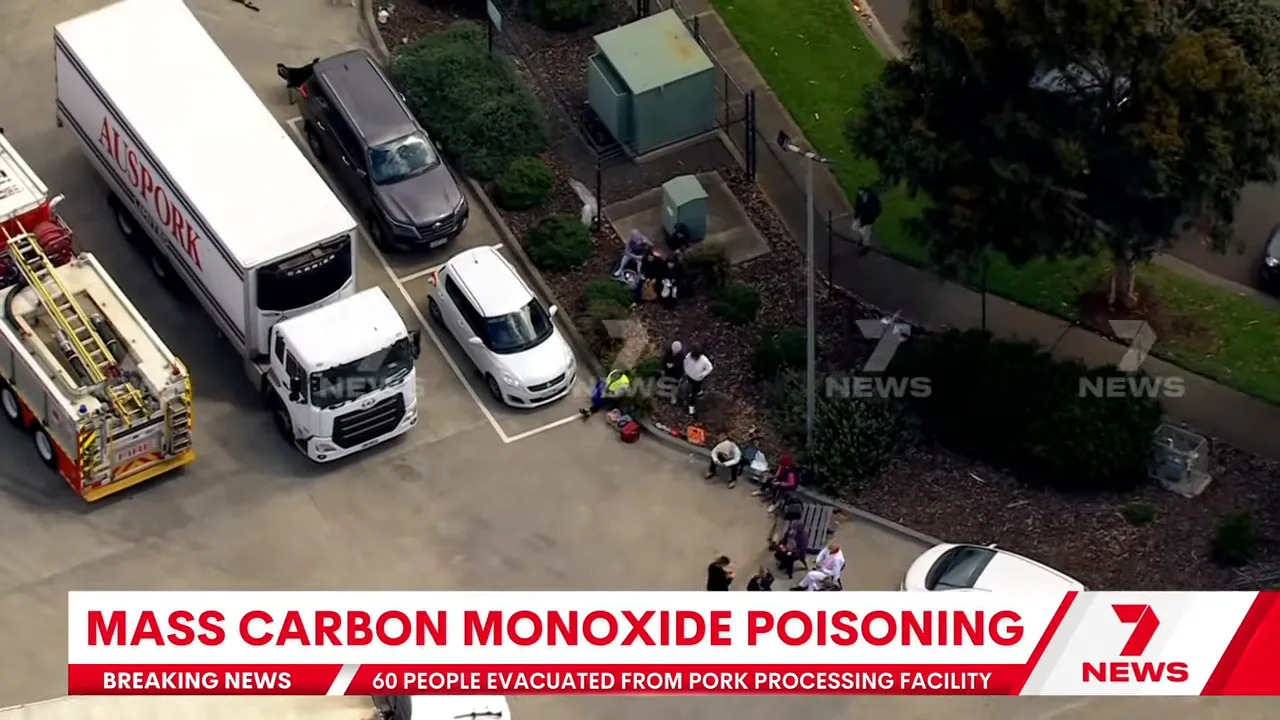 Close aerial view of evacuated workers seated outside a meat‑processing plant with emergency responders and medical equipment nearby.