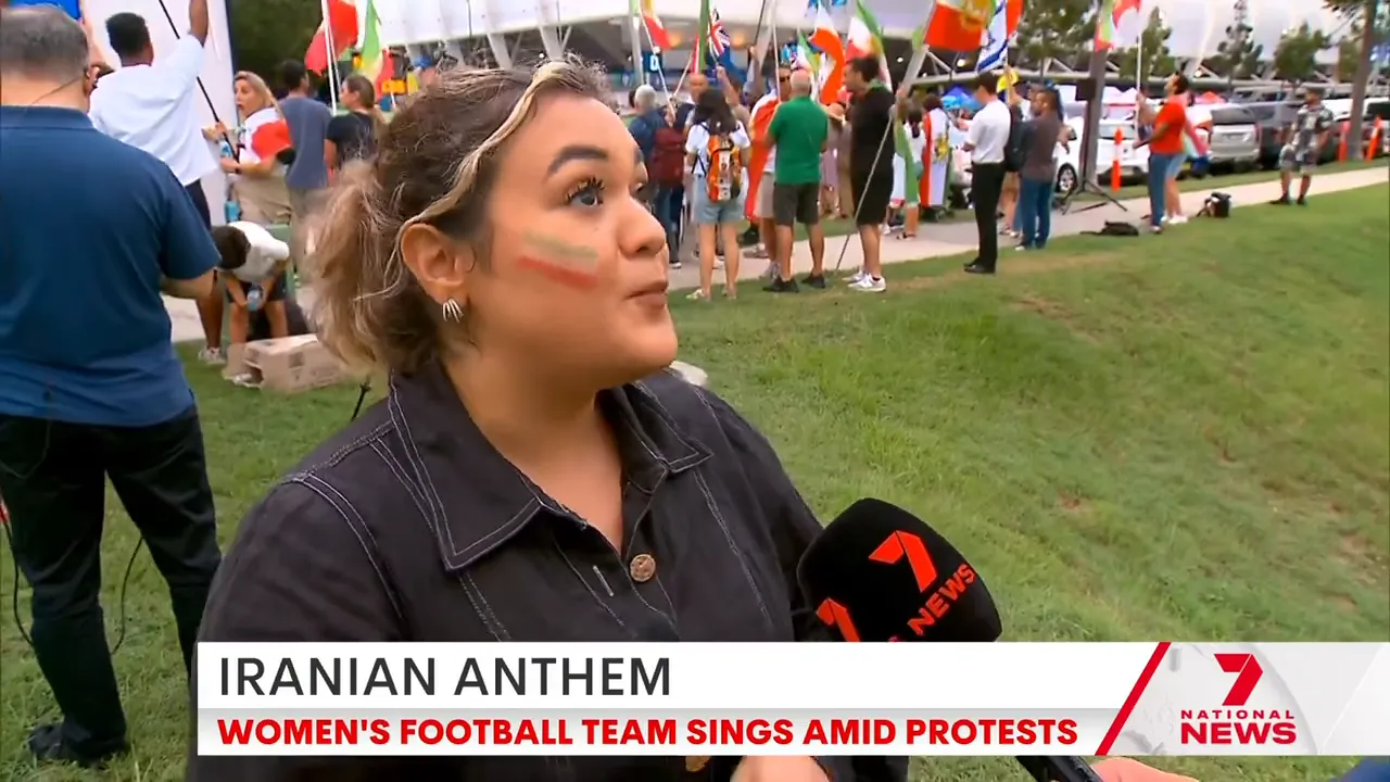 Wide shot of a protester with Iranian-flag face paint speaking to a Seven News microphone with a crowd of demonstrators and flags behind her