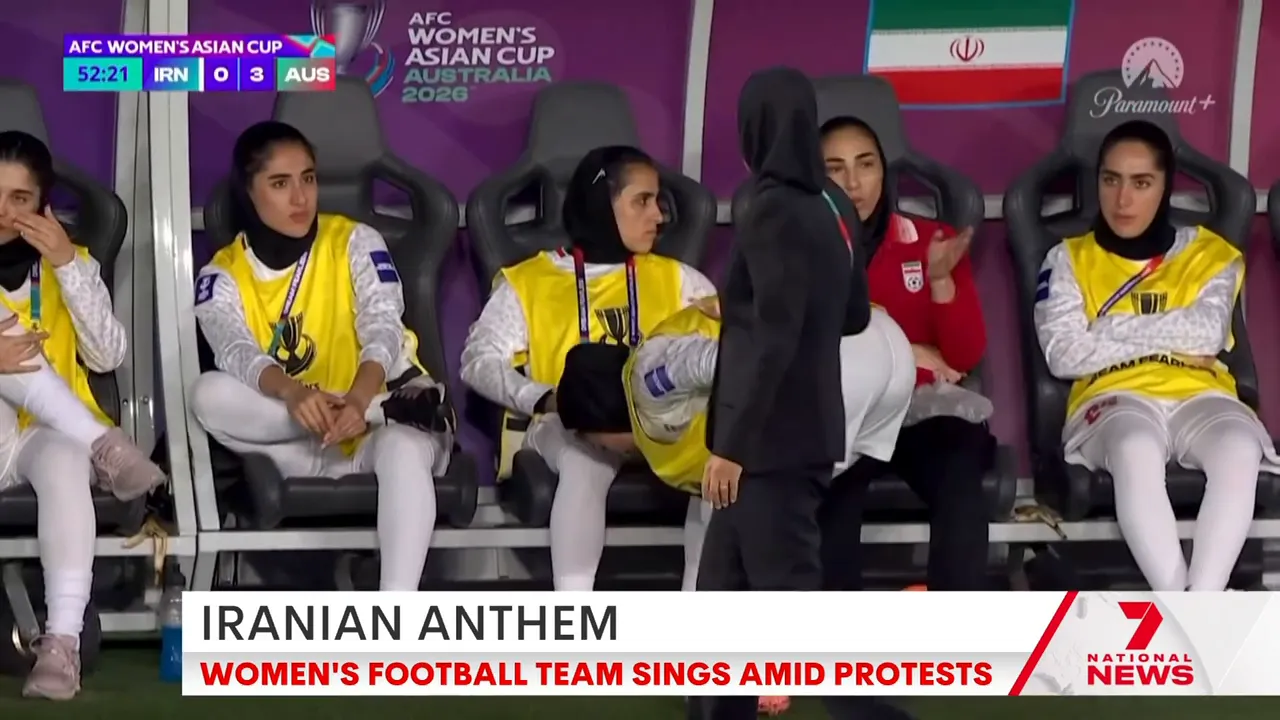 Iranian women's team players seated on the substitutes' bench wearing hijabs and training bibs, Iranian flag visible behind them.