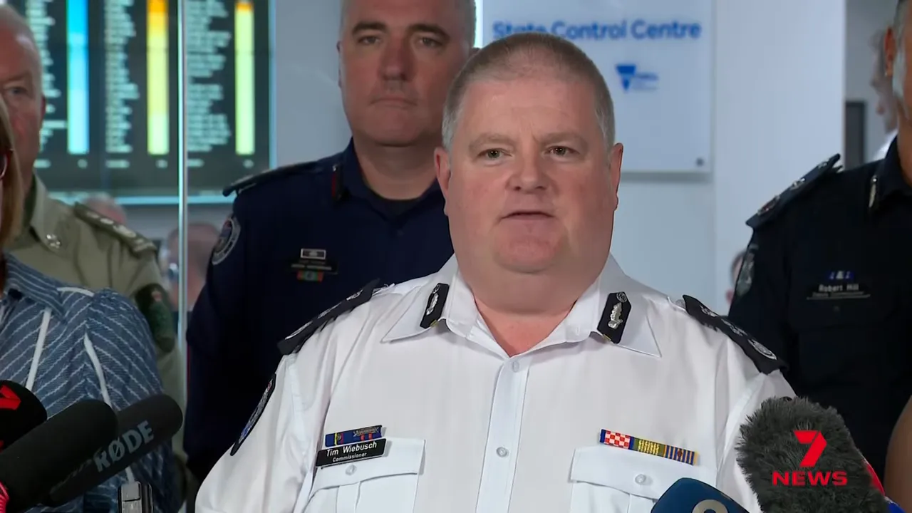 Emergency services official facing camera at a press briefing, microphones and news branding in frame.