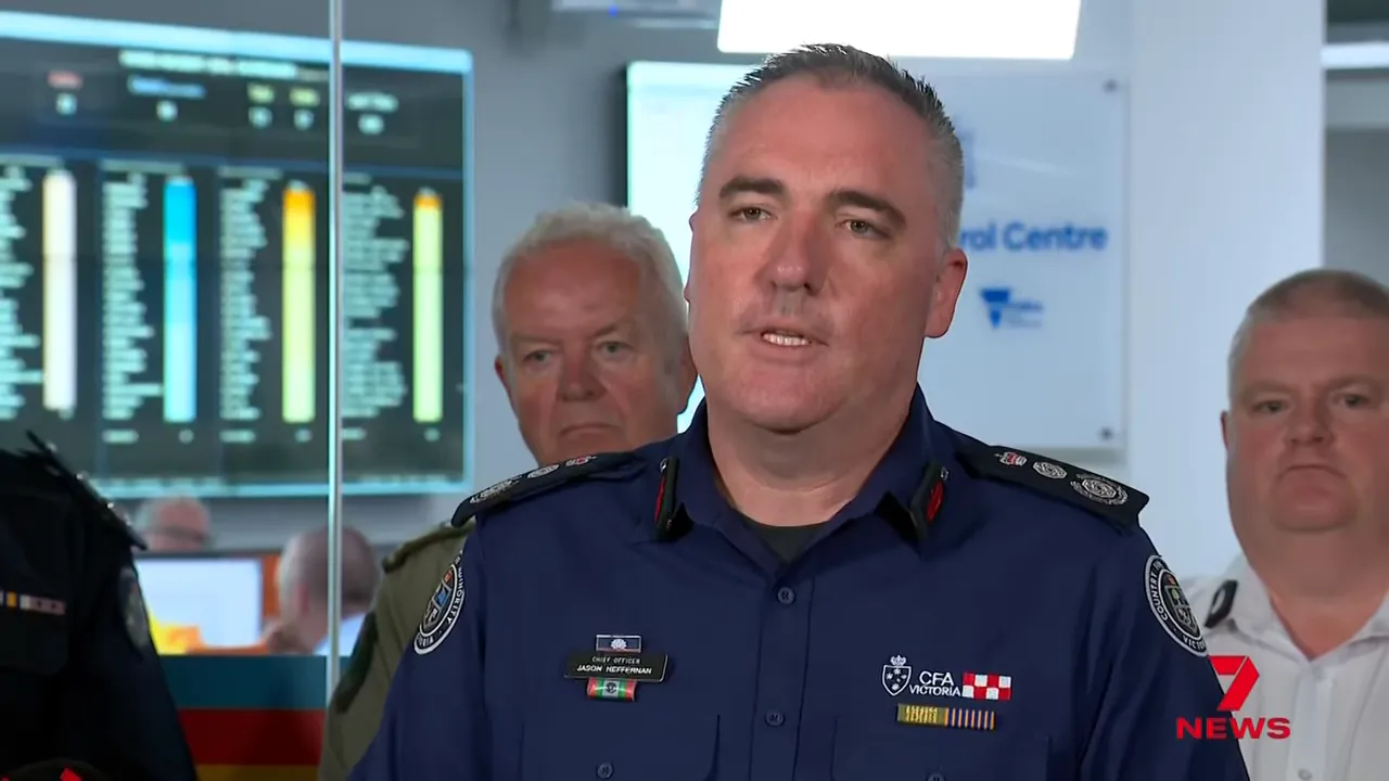 Emergency service chief speaking at a press briefing in an operations centre, with monitoring screens and other officials in the background.