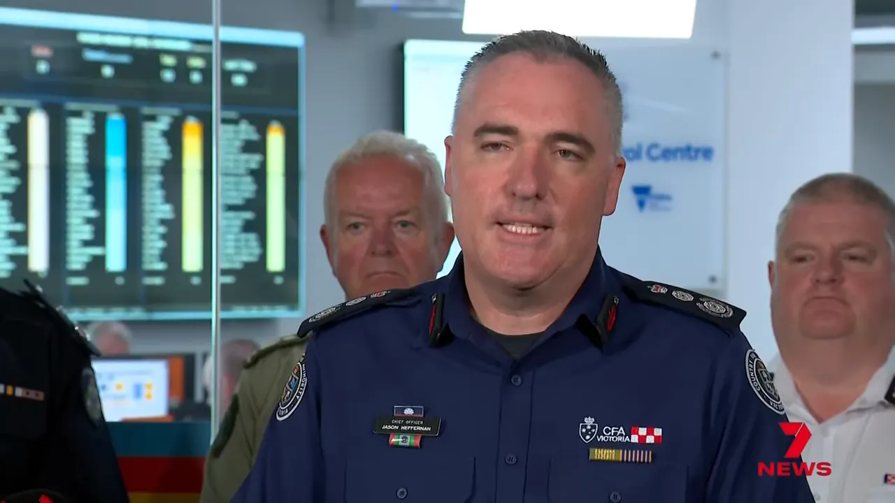 Fire authority spokesperson speaking at a press conference with emergency operations displays in the background