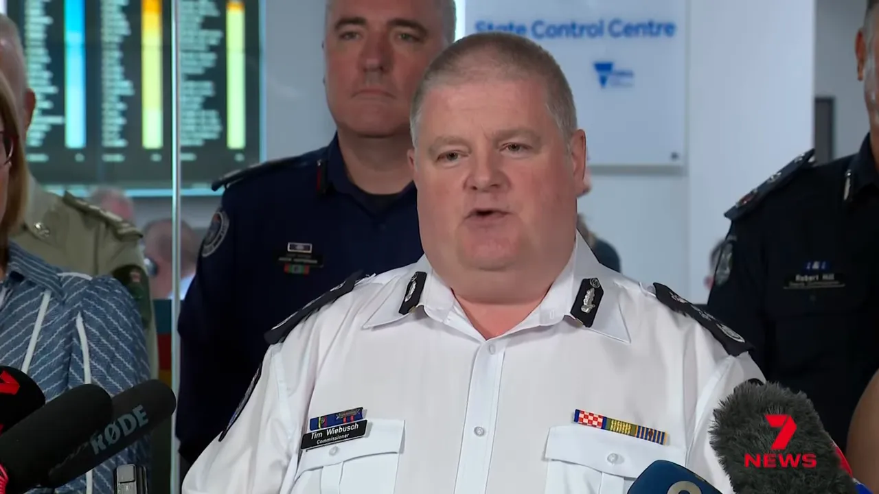 Emergency services officer speaking at a press conference with microphones and command staff behind him.
