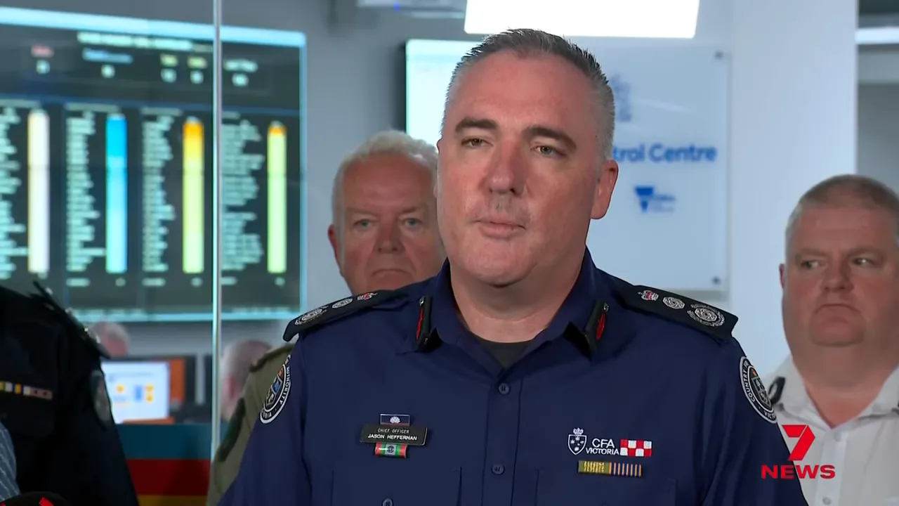 Senior emergency services official speaking at a press briefing in a control centre with monitoring screens behind