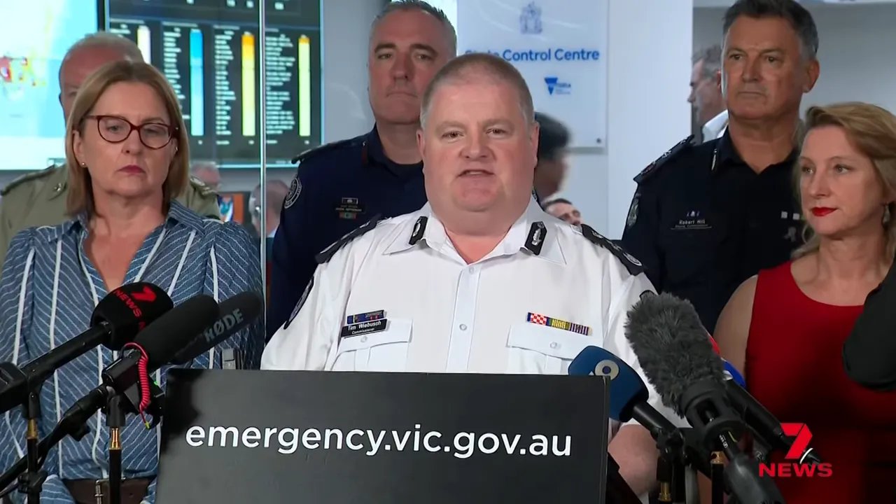 State emergency press conference showing officials, microphones and 'emergency.vic.gov.au' sign