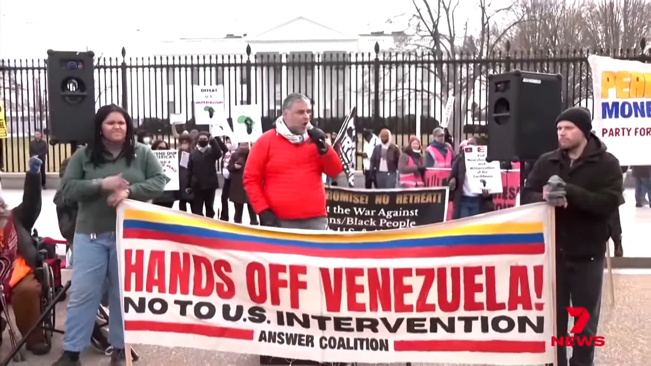Wide shot of a crowd in front of the White House fence holding a large 'Hands off Venezuela' banner opposing US intervention.