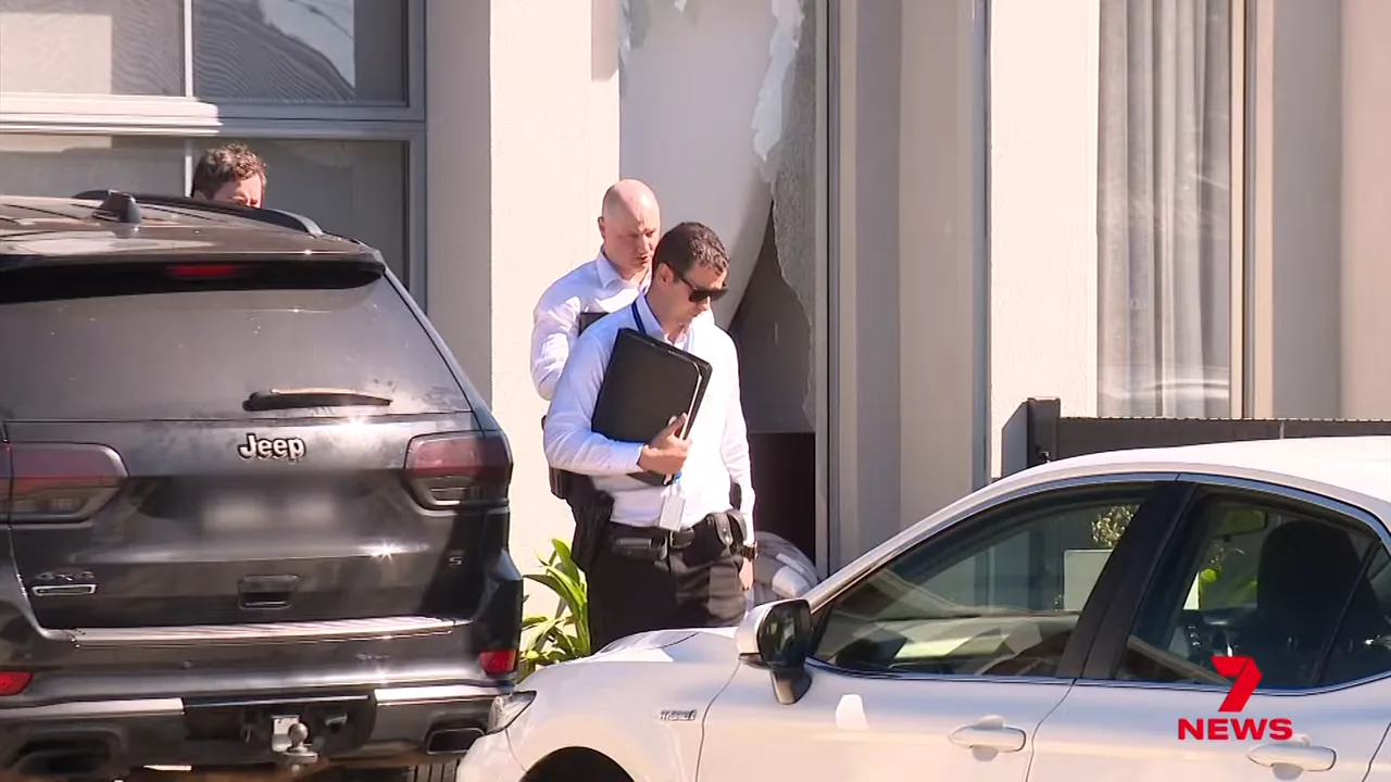 Police investigators at a house entrance with a damaged window and parked cars
