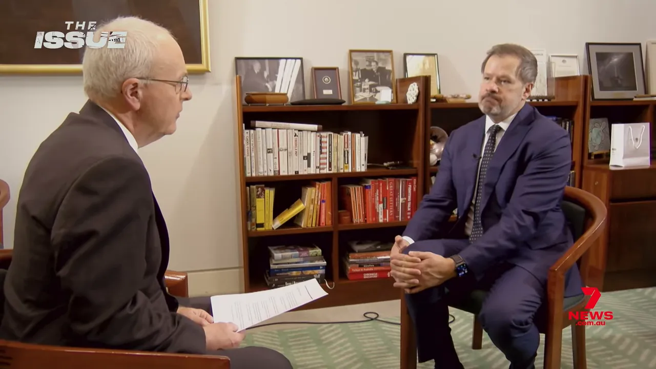 Wide shot of an in‑office interview showing an MP seated across from an interviewer with a bookcase behind them.