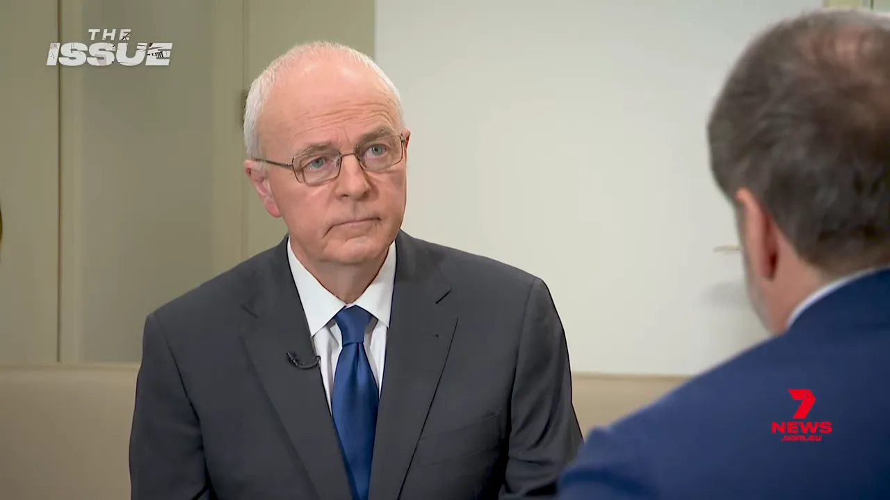 Wide mid-shot of an interviewer listening attentively to the interviewee (seen from behind) in a studio interview setting.
