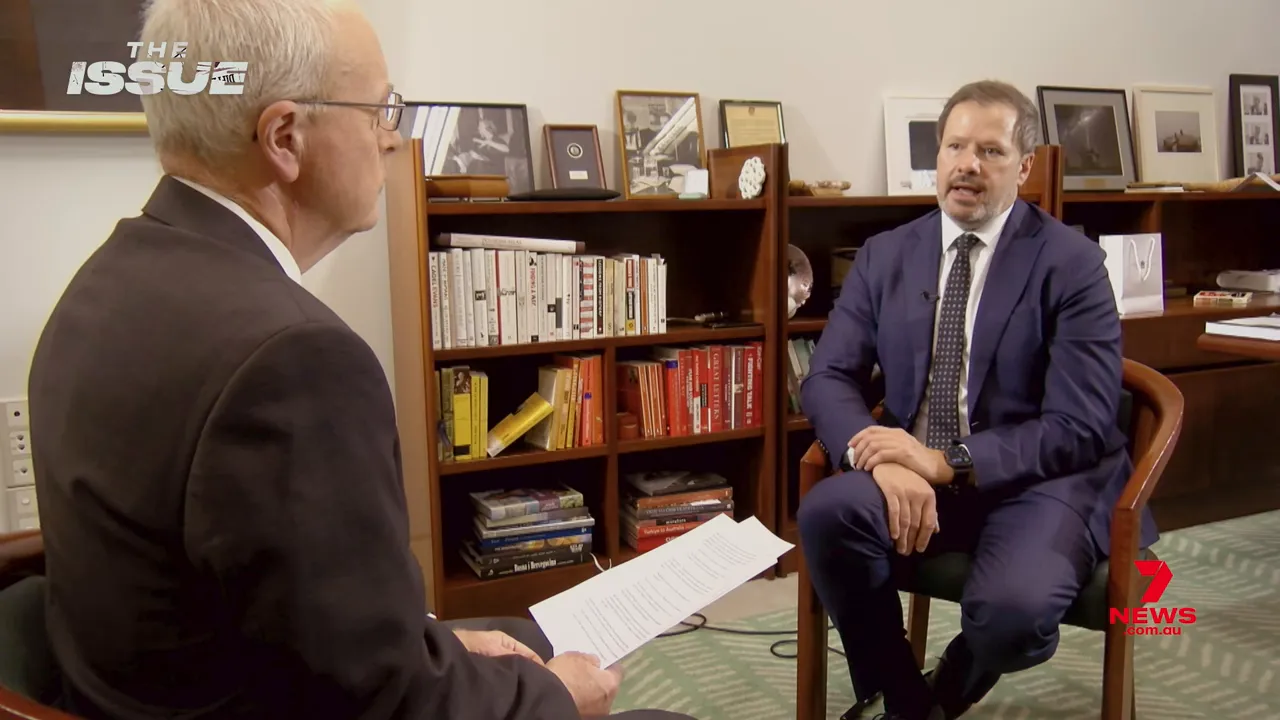 Two‑shot of an in‑office televised interview showing the MP and interviewer with bookshelves behind them
