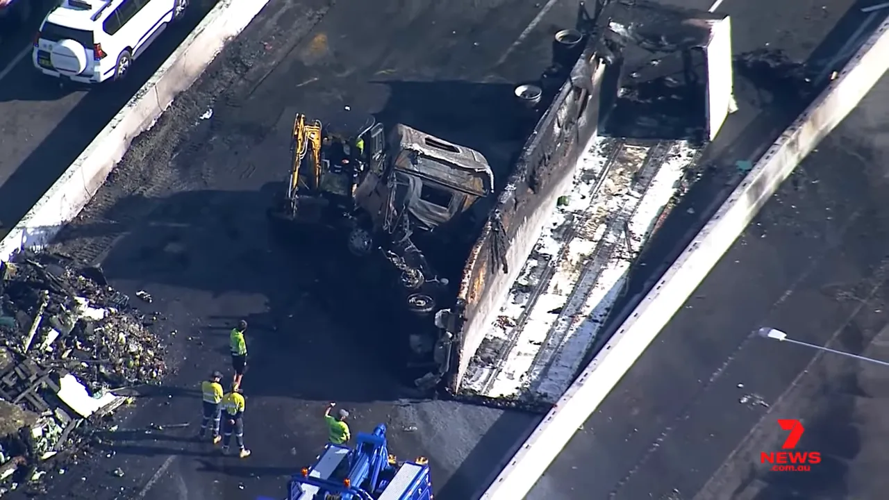 Aerial image of a burned semi-trailer on its side with a charred cab, debris and recovery crew on the motorway