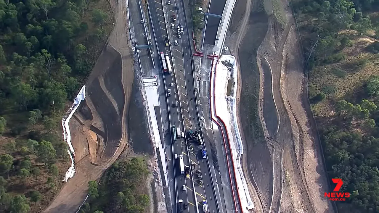 Wide aerial view of the M7 crash scene showing emergency vehicles, the burned trailer and white containment booms along the embankment.