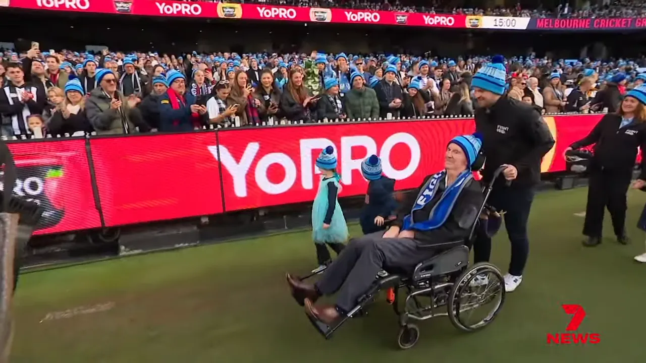 Stadium crowd wearing blue beanies with a person in a wheelchair being pushed along the sideline