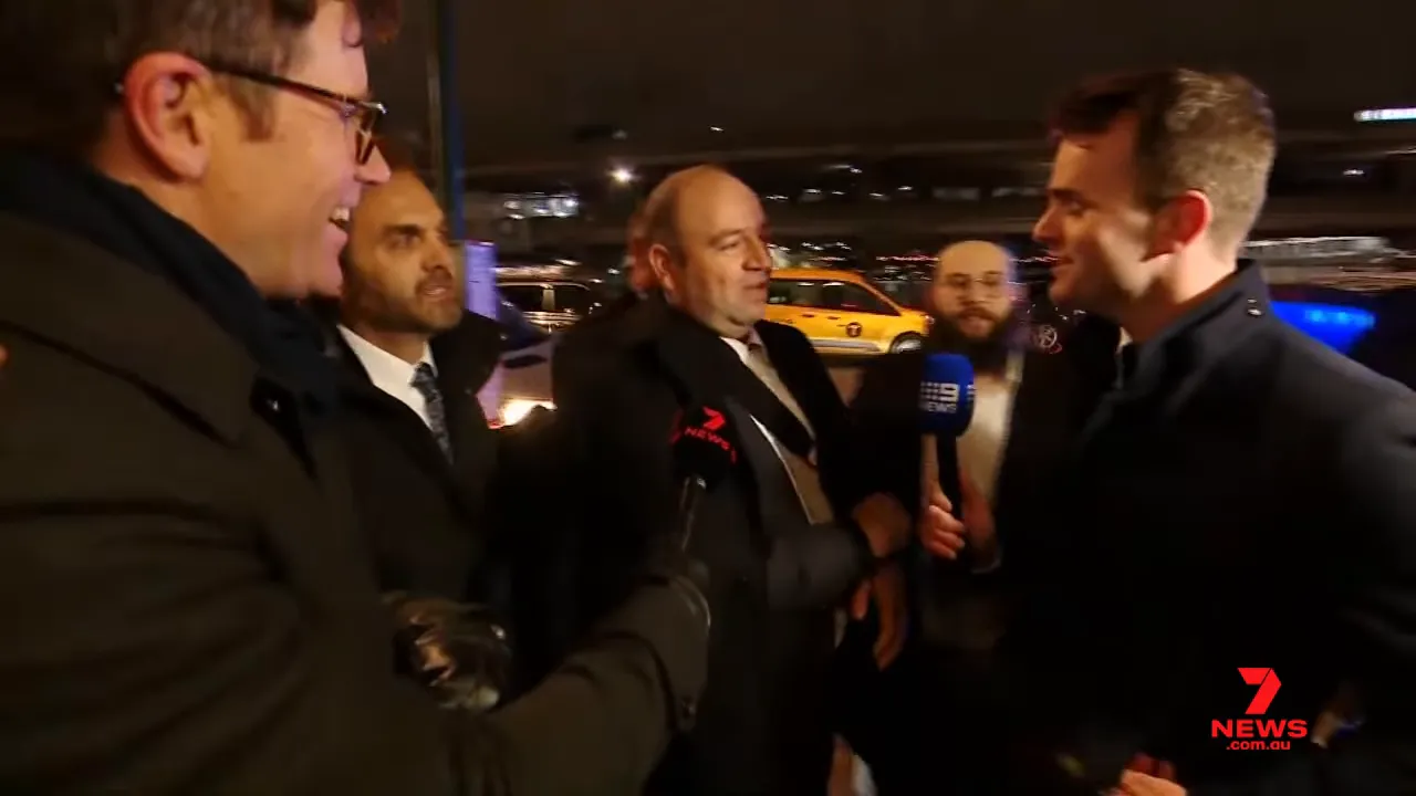 Man speaking with reporters and supporters at a nighttime arrival, microphones and a taxi in the background