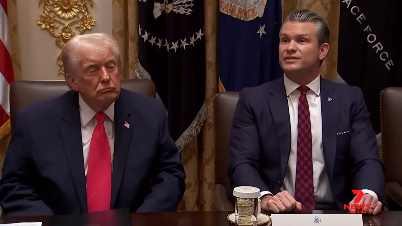 Two seated officials at a conference table in a formal room, both visible and clearly focused, one preparing to speak.