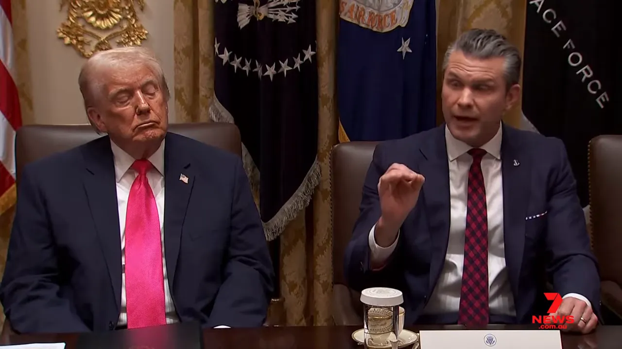 Two officials seated at a conference table during a press briefing, one making a clear hand gesture while speaking