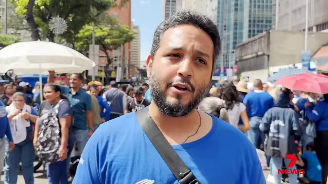 Man speaking to camera in a crowded Caracas street protest, with demonstrators, umbrellas and city buildings visible behind him.