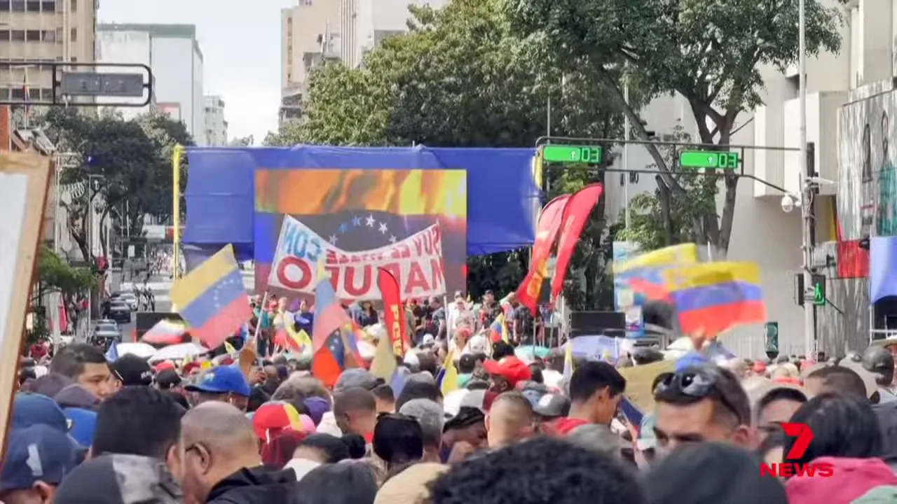 Large daytime street demonstration with a stage, Venezuelan flags and a dense crowd of supporters.