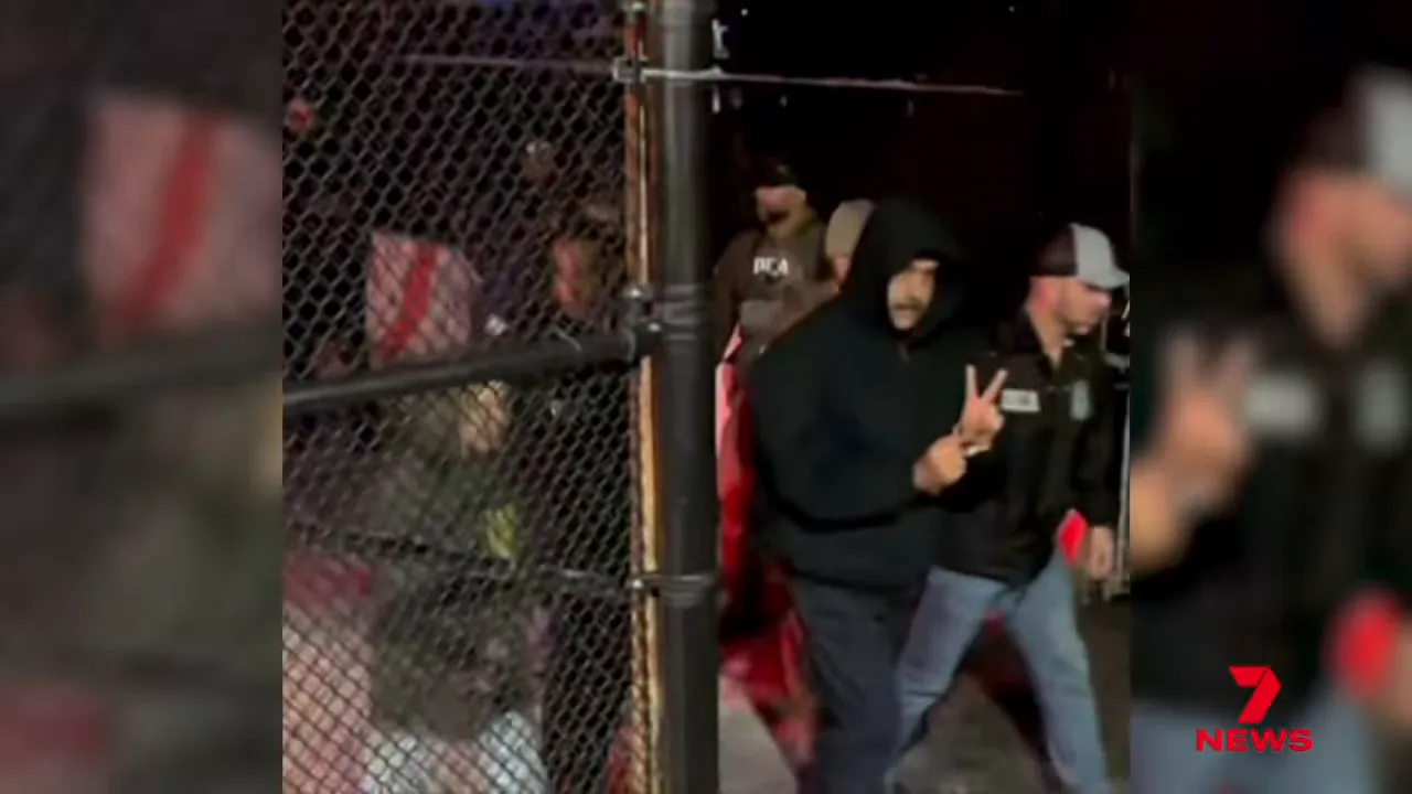 Hooded man making a hand gesture while being escorted past a chain‑link fence by law‑enforcement agents at night.