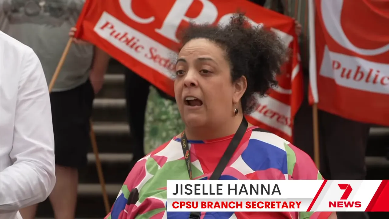 Union members holding red 'Public Sector' flags while a union representative speaks at a press event.