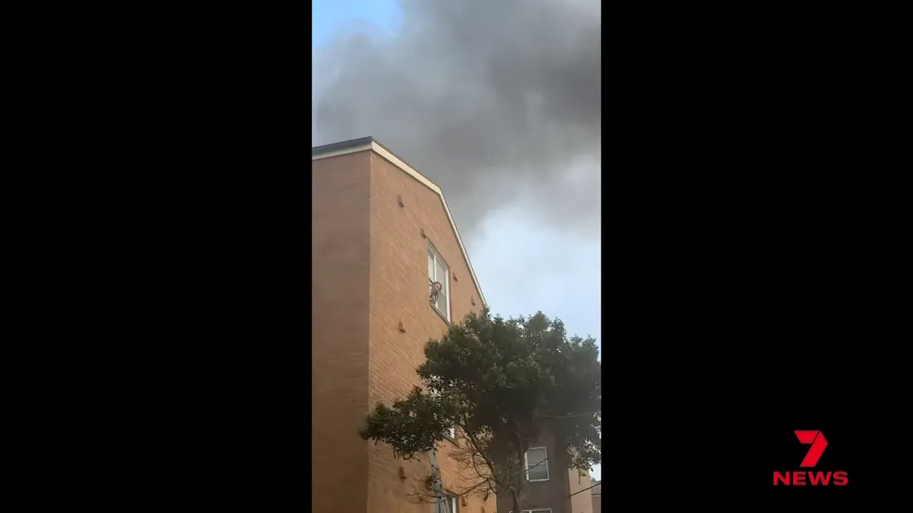 Thick smoke above a three-storey apartment with a ladder leaned toward an upper-floor window