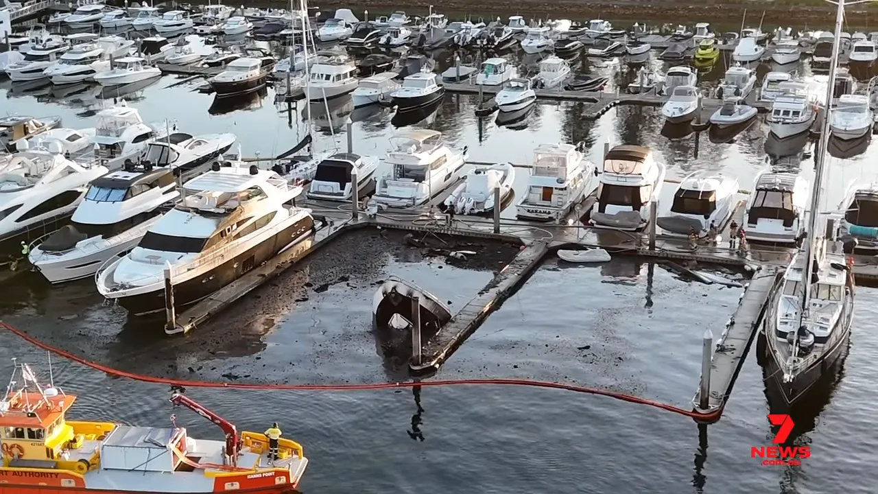 High-resolution aerial image of a sunken, charred yacht in a marina with containment booms and a rescue/salvage vessel alongside
