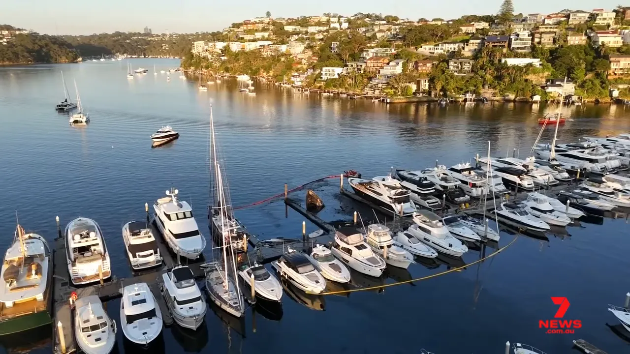 Daylight aerial view of Spit Bridge Marina showing yellow containment booms around berths and a partially sunken yacht after the blaze.