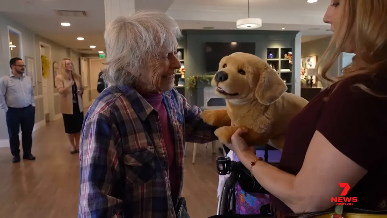 elderly woman smiling while touching a lifelike robotic puppy held by a caregiver in a care home