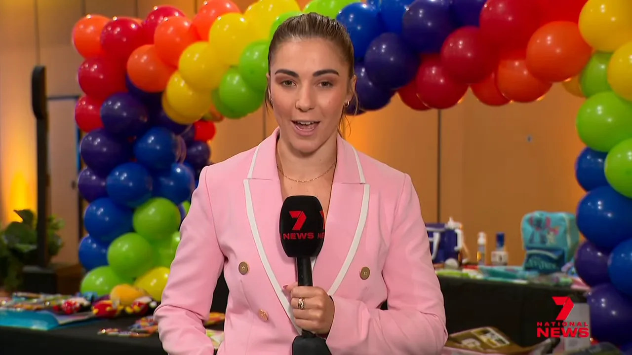 Wide shot of a 7NEWS reporter holding a microphone with a rainbow balloon arch and showbag table visible in the background