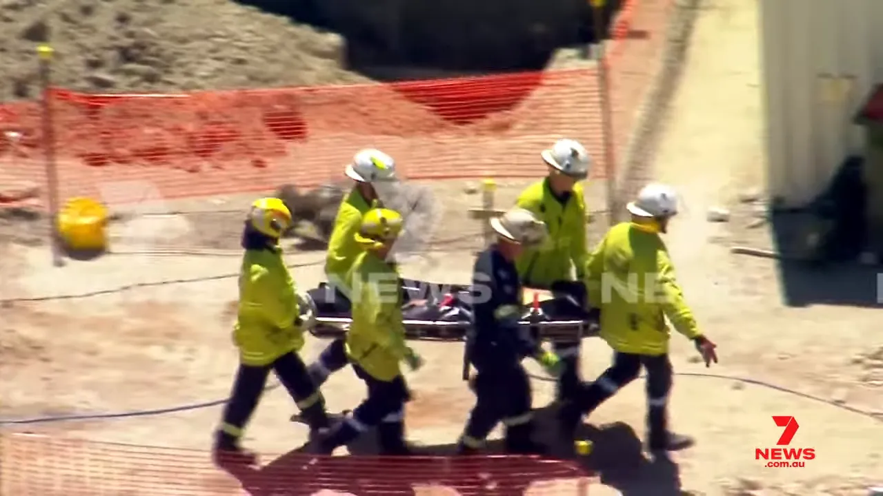 Firefighters and paramedics carrying an injured worker on a stretcher across the construction site toward an ambulance.