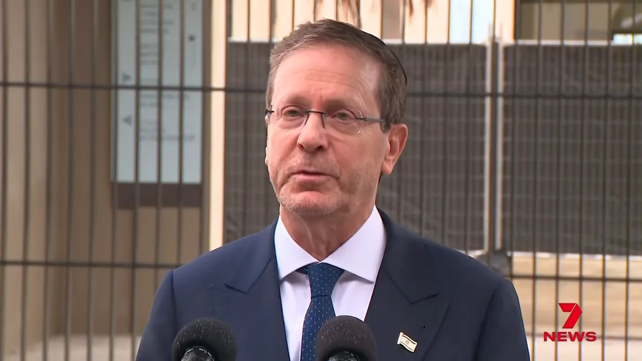 Clear frontal shot of a man in a suit speaking at an outdoor memorial with two press microphones in the foreground.