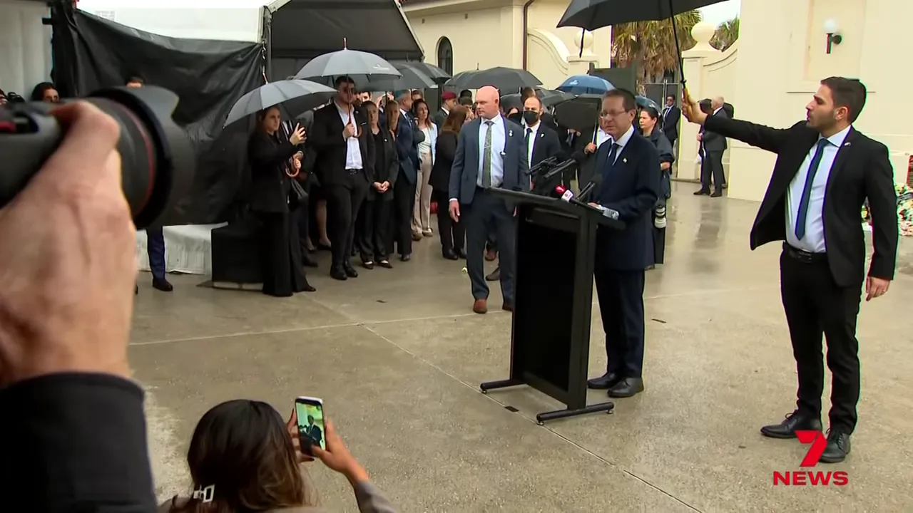 Wide view of the Bondi memorial ceremony showing the speaker at a podium, an aide holding an umbrella, and attendees gathered under umbrellas.