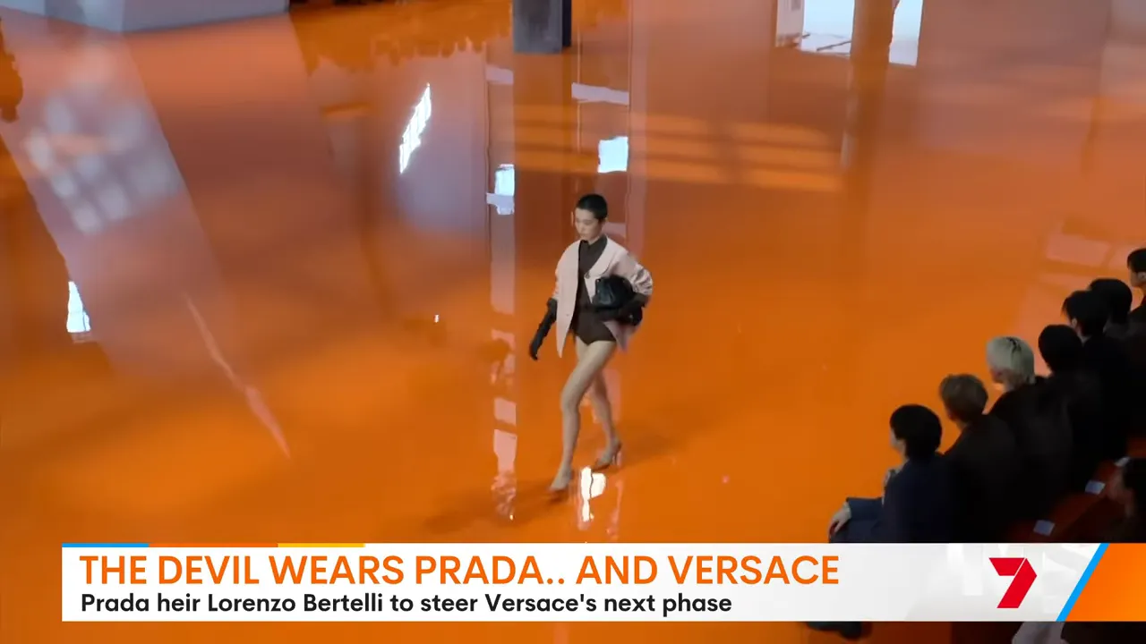 Versace model walking on a reflective orange catwalk with audience visible at the edge